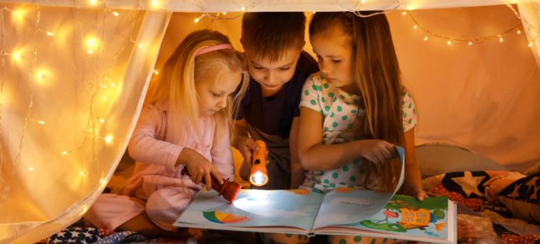 Tres niños leyendo un libro dentro de un fuerte de mantas, participando en desafíos de lectura de verano para transformar la lectura en casa en una aventura emocionante.